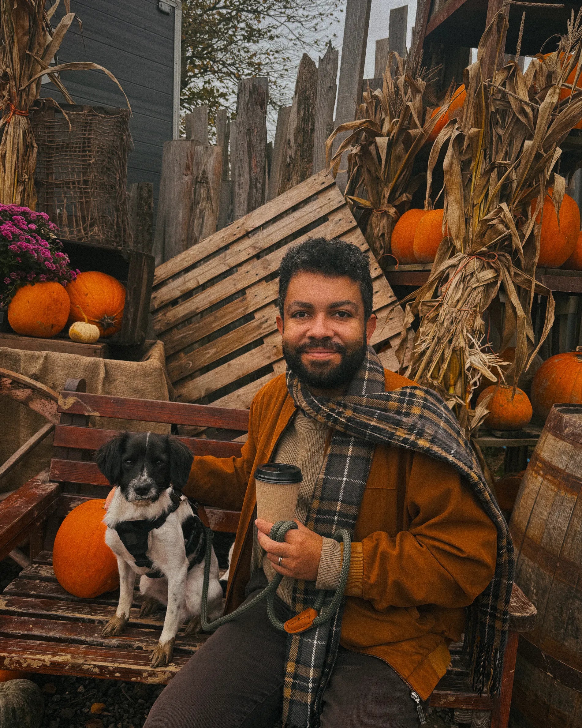 Artie sitting on a bench next to Leslie holding a coffee cup in an autumnal setting with pumpkins and corn stalks.